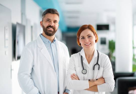 Ein Arzt und eine Ärztin stehen selbstbewusst lächelnd in einem Krankenhaus